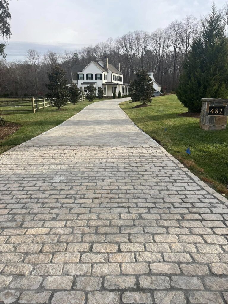 A long, light stone driveway leads to a white two-story house with dark shutters. The lawn is green and well-kept, framed by trees and bushes. A mailbox with the number 482 stands on the right near the driveway entrance. - Hutchins Landscaping and Lawn Maintenance Rouguemont, NC