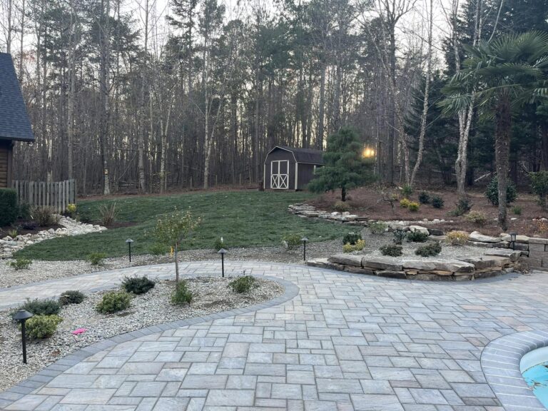 A neatly landscaped backyard features a stone patio with garden beds of shrubs and small plants bordered by rocks. Beyond the patio, there is a grassy area and a brown wooden shed. Tall trees and bushes line the background, creating a woodland setting. - Hutchins Landscaping and Lawn Maintenance Rouguemont, NC