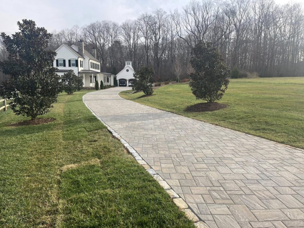 A paved stone driveway curves through a large, green lawn toward a white house with black shutters and an attached garage. Leafless trees line the background, and a few small trees are planted along the driveway. The sky is slightly overcast. - Hutchins Landscaping and Lawn Maintenance Rouguemont, NC
