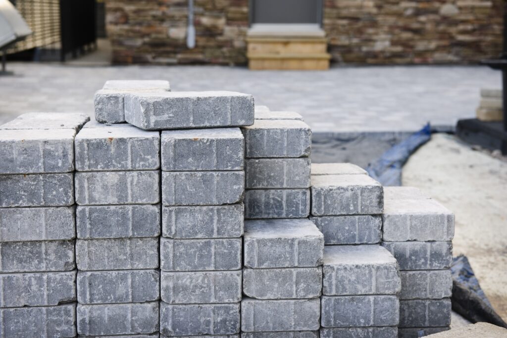A stack of gray rectangular concrete pavers sits on a Rougemont construction site. The pavers are neatly arranged, with sand and partially paved ground nearby. In the background, a patio area and brick wall highlight Durham landscaping work. - Hutchins Landscaping and Lawn Maintenance Rouguemont, NC