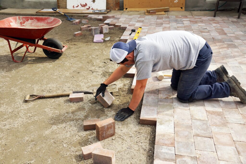 A person wearing gloves, a gray shirt, blue pants, and a white cap kneels on the ground in Durham, placing rectangular paving stones into a pattern as part of landscaping work. A shovel, wheelbarrow, and scattered bricks lie nearby on the sandy surface. - Hutchins Landscaping and Lawn Maintenance Rouguemont, NC