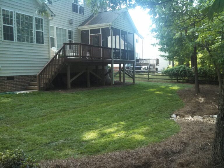A backyard in Rougemont with a screen porch and wooden stairs leading to a neatly landscaped lawn. The grass is mowed, bordered by pine straw and mulch, with trees, shrubs, and dappled sunlight creating a peaceful Durham retreat. - Hutchins Landscaping and Lawn Maintenance Rouguemont, NC