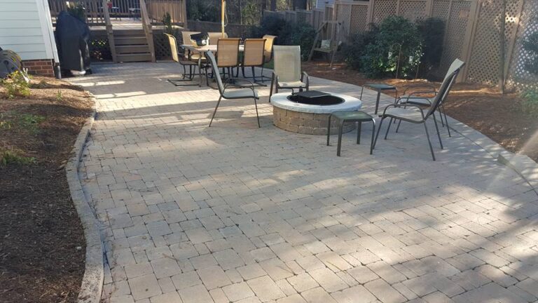 Outdoor patio with light stone pavers, a round fire pit surrounded by metal chairs, and a long dining table in the background. Landscaping features like shrubs and mulch line the wood fencing and lattice panels of this Durham or Rougemont retreat. - Hutchins Landscaping and Lawn Maintenance Rouguemont, NC